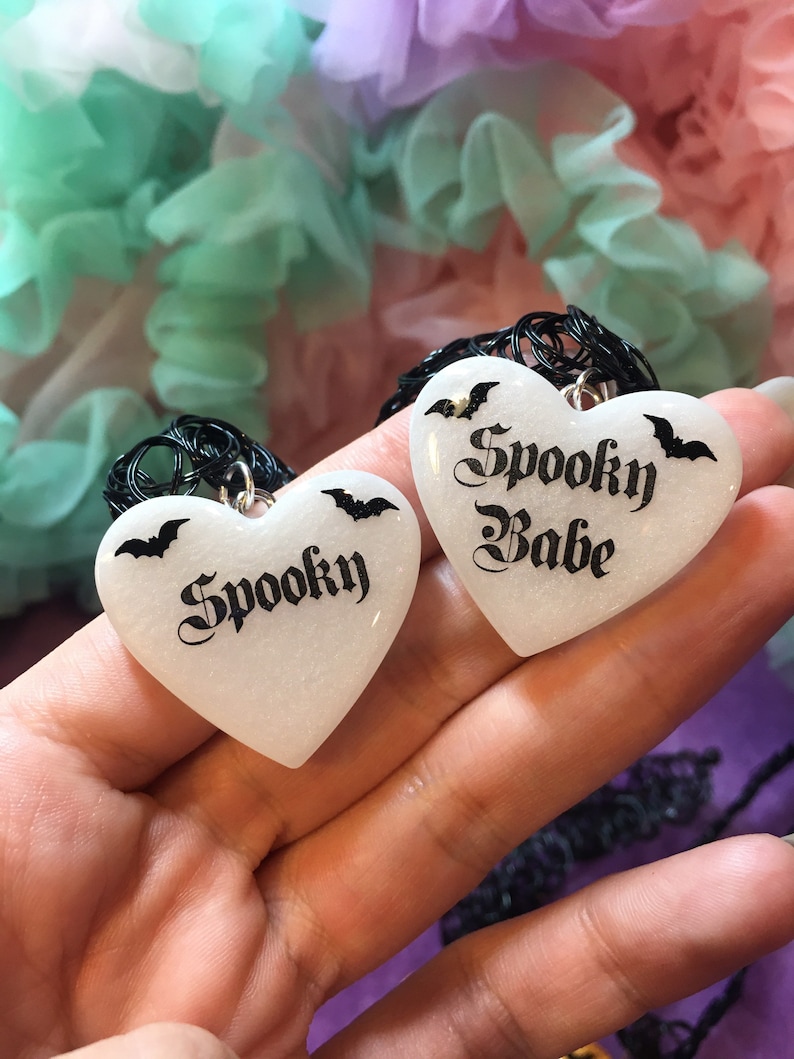 May include: Two white heart-shaped pendants with black bats and the words "Spooky" and "Spooky Babe" in black script.