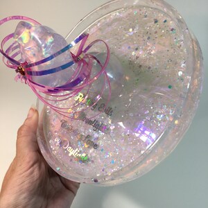May include: A clear, round, glitter-filled resin tray with a pink and purple ribbon bow. The tray has the text "Riley & Edith Moonlight  Creating a Love  by Daylight" inscribed on the bottom.