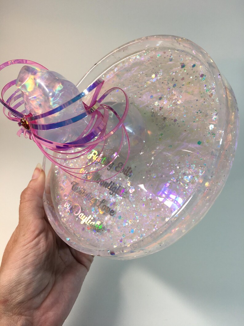 May include: A clear, round, glitter-filled resin tray with a pink and purple ribbon bow. The tray has the text "Riley & Edith Moonlight  Creating a Love  by Daylight" inscribed on the bottom.