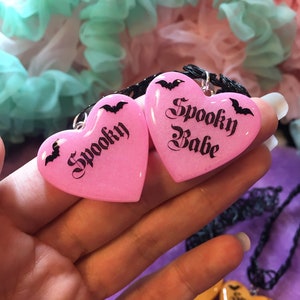May include: Two pink heart-shaped pendants on a black chain necklace. The pendants are made of resin and have a glittery finish. The left pendant says "Spooky" and the right pendant says "Spooky Babe".