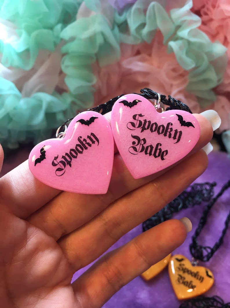 May include: Two pink heart-shaped pendants on a black chain necklace. The pendants are made of resin and have a glittery finish. The left pendant says "Spooky" and the right pendant says "Spooky Babe".