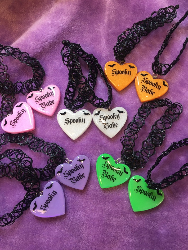 May include: A set of eight black choker necklaces with heart-shaped pendants. The pendants are made of clear plastic and have different colors and glitter. Each pendant has the words "Spooky Babe" written on it in black lettering. The pendants also have a small black bat design.
