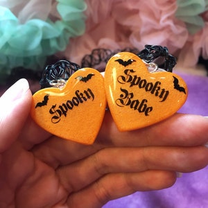 May include: Two orange heart-shaped pendants with black bats and the words "Spooky" and "Spooky Babe" written in black script.