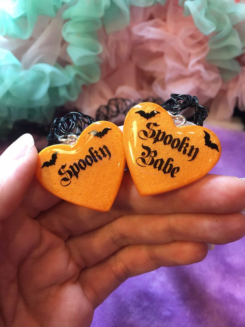 May include: Two orange heart-shaped pendants with black bats and the words "Spooky" and "Spooky Babe" written in black script.