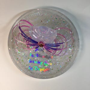 May include: A clear round resin coaster with iridescent glitter and a pink and purple resin flower with a gold accent. The coaster has the text "Giving Love by Moonlight, Receiving Love by Daylight" in rainbow iridescent lettering.