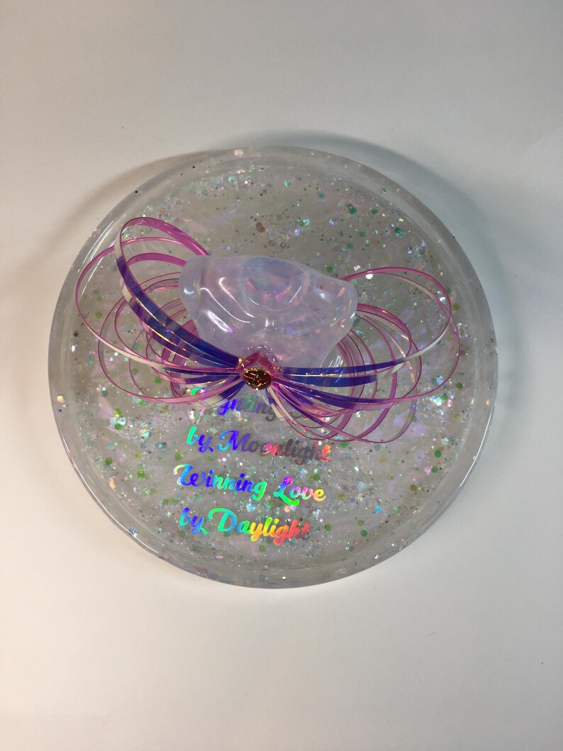 May include: A clear round resin coaster with iridescent glitter and a pink and purple resin flower with a gold accent. The coaster has the text "Giving Love by Moonlight, Receiving Love by Daylight" in rainbow iridescent lettering.