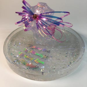 May include: A clear round tray with iridescent glitter and the words "Fighting Evil with Moonlight" and "Uniting Love with Starlight" in iridescent lettering. A large iridescent pink bow sits on top of the tray.