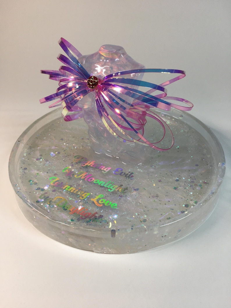 May include: A clear round tray with iridescent glitter and the words "Fighting Evil with Moonlight" and "Uniting Love with Starlight" in iridescent lettering. A large iridescent pink bow sits on top of the tray.