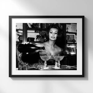 Könnte beinhalten: Schwarz-Weiß-Druck in einem Rahmen, der eine Frau zeigt, die Champagner in zwei Gläser gießt. Das Bild ist eine Vintage-Fotografie, wahrscheinlich aus der Mitte des 20. Jahrhunderts, und wird an einer weißen Wand ausgestellt. Die Frau ist der Mittelpunkt, mit einer Flasche in der Hand.