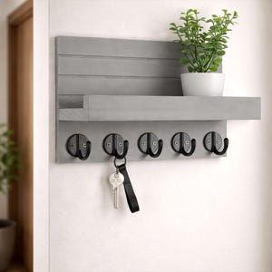Puede incluir: Un llavero de pared de madera gris con un estante y cinco ganchos negros. Una pequeña planta en maceta se encuentra en el estante. Las llaves cuelgan de un gancho. El llavero está montado en una pared blanca.