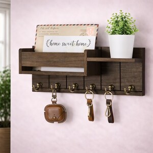 Può includere: Un organizer da parete in legno marrone scuro con un ripiano, una fessura per la posta e ganci per chiavi. L'organizer contiene una piccola pianta in vaso, una busta e una carta con la scritta "home sweet home". Diverse chiavi e una custodia AirPod marrone sono appese ai ganci.