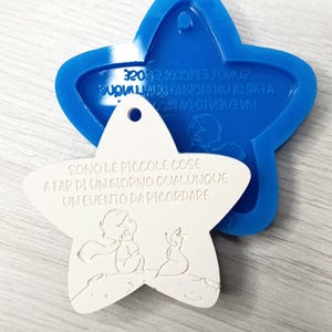 Può includere: Un ciondolo bianco a forma di stella con uno stampo blu a forma di stella. Il ciondolo presenta un'illustrazione di un personaggio e una volpe, con testo in italiano. Lo stampo blu ha anche testo in italiano. Il ciondolo ha un foro per appenderlo.