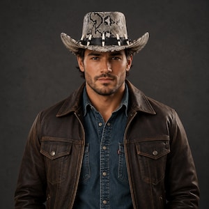 Könnte beinhalten: Ein Cowboyhut mit Schlangenmuster, verziert mit schwarzem Band und weißen Akzenten. Der Hut hat eine breite Krempe und eine hohe Krone. Er wird mit einer braunen Lederjacke und einem Jeanshemd getragen.