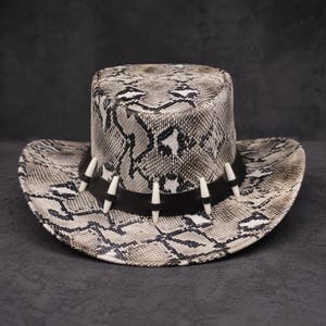 May include: A snakeskin-effect cowboy hat with a black band and white embellishments. The hat has a wide brim and a structured crown. The snakeskin design uses grey, black, and white, giving a textured look. This hat is a fashionable accessory.