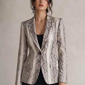 May include: A snakeskin-patterned blazer in neutral tones of beige, gray, and black. The tailored jacket features a single-button closure, notched lapels, and two front pockets. The model is wearing a black top and black pants.
