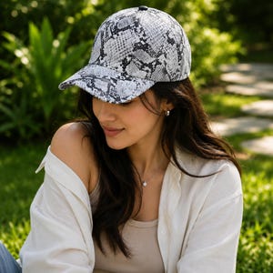 May include: A baseball cap with a snakeskin pattern in shades of gray and white. The cap has a curved brim and a black button on top. The hat is worn by a person.