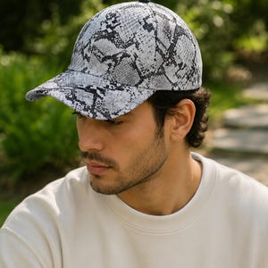 May include: A baseball cap with a snakeskin pattern in shades of gray and black. The cap has a curved brim and a black button on top. The hat is worn by a person wearing a cream-colored sweatshirt.