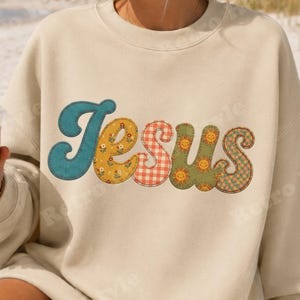Könnte beinhalten: Ein beigefarbenes Sweatshirt mit Rundhalsausschnitt und dem Wort Jesus in großen, stilisierten Buchstaben. Jeder Buchstabe hat ein einzigartiges Muster: blaugrüner Kord, gelbe Blumen, rot-weißes Vichy-Karo, olivgrün mit lächelnden Sonnen und grüne Blumen.