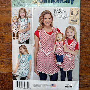 May include: Simplicity sewing pattern J0204 for 1920s vintage aprons. The pattern features multiple views of aprons with a red and white patterned design. The package includes images of women and girls wearing the aprons. The pattern is designed and printed in the USA.