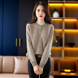 May include: A light brown cardigan with a round neckline and button-up closure. The long-sleeved sweater is paired with black trousers and a gold-tone belt. The woman is wearing earrings and has red lipstick.