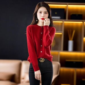 May include: A red button-down cardigan with long sleeves. The cardigan has silver buttons and is paired with black pants. A gold belt buckle is visible. The background includes a dark wall and a wooden shelf.