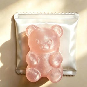 May include: A translucent, light pink, bear-shaped object with a smooth, glossy finish, positioned directly in front of a square, silver-colored metallic foil packet with a crimped top and bottom edge, featuring a simple, molded facial design.