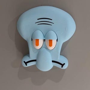Può includere: Una scultura da parete azzurra a forma di testa di personaggio dei cartoni animati con una fronte ampia, palpebre pesanti, pupille arancioni e un naso prominente. Il personaggio presenta linee nere per le sopracciglia e una bocca rivolta verso il basso.