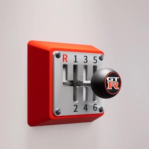 May include: A wall-mounted red gear shifter display featuring a silver metal gate with black engraved numbers 1, 2, 3, 4, 5, 6 and the letter R. A black spherical shift knob with a red and white GT-R logo is attached to the center metal lever.