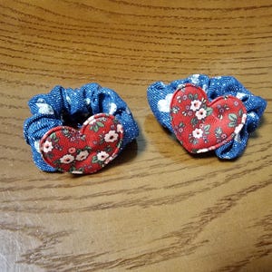 Può includere: Due elastici per capelli blu con un motivo floreale bianco, ciascuno con un'applicazione a forma di cuore rosso. I cuori sono decorati con piccoli fiori bianchi e foglie verdi, creando un'estetica affascinante e fatta a mano.