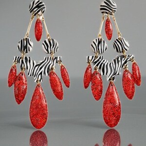 Peut inclure: Paire de boucles d'oreilles pendantes avec un motif zébré noir et blanc. Elles comportent des éléments circulaires et rectangulaires, des accents dorés et des pendentifs rouges scintillants en forme de goutte.
