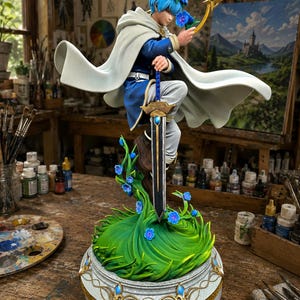Puede incluir: Figura detallada de un personaje con un atuendo azul y blanco, sosteniendo una espada y una flor. La figura está adornada con una corona floral azul y una capa blanca fluida. La base presenta follaje verde y flores azules, sobre una plataforma decorativa con ribete dorado.