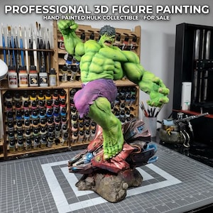 以下が含まれることがあります： 手描きのハルクのコレクターズフィギュア。緑色の肌と紫色のショートパンツで、ダイナミックなポーズをとっています。フィギュアは、赤、青、茶色のトーンが混ざった詳細なベースの上に立っています。背景には絵画用品が写っています。