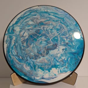 May include: Axiom Discs disc golf disc with a blue and white marbled design. The disc has a black rim and a silver graphic in the center. The disc is displayed on a wooden stand.