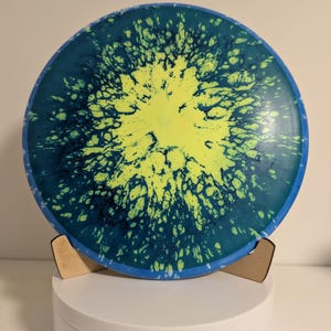 May include: A disc golf disc with a vibrant, eye-catching design. The disc features a bright yellow center that splatters outwards into a dark teal and blue pattern. The disc is supported by a wooden stand, showcasing its unique design.