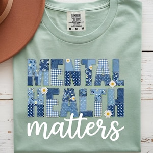 May include: A sage green t-shirt featuring the text MENTAL HEALTH in a blue patchwork quilt pattern with small white daisy accents, and the word matters in white script below. A brown felt hat is positioned to the left of the t-shirt.