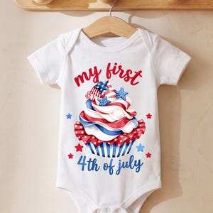 May include: White baby bodysuit with a patriotic cupcake design. The cupcake features red, white, and blue frosting, stars, and the text "my first 4th of July". The bodysuit is hanging on a wooden hanger.