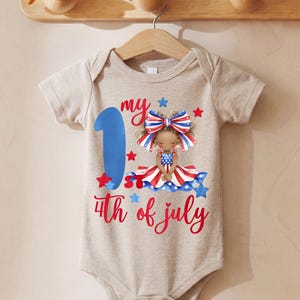May include: A beige baby bodysuit with a graphic celebrating the 4th of July. The design includes a large blue number 1, the words "my 4th of July" in red script, and a cartoon girl in a patriotic outfit. The bodysuit is hanging on a wooden hanger.