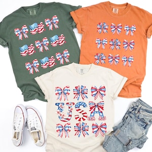 May include: Three t-shirts in olive green, orange, and cream, each adorned with patriotic bow designs in red, white, and blue. A pair of white sneakers and a pair of blue jeans are also in the image. The cream shirt has the word "USA" in the design.