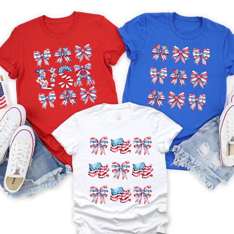 May include: Three t-shirts in red, white, and blue, each featuring a patriotic bow design. The red shirt has the word "USA" in a bow design. The white shirt has American flag bow designs. The blue shirt has bow designs.