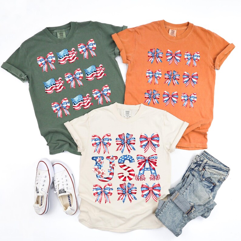 May include: Three t-shirts in olive green, orange, and cream, each adorned with patriotic bow designs in red, white, and blue. A pair of white sneakers and a pair of blue jeans are also in the image. The cream shirt has the word "USA" in the design.