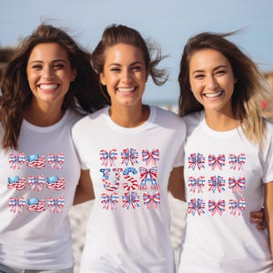 May include: Three women wearing white t-shirts with patriotic bow designs in red, white, and blue. The middle shirt has the word "USA" in a similar bow design. The shirts are perfect for celebrating the 4th of July.