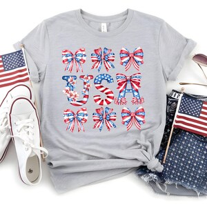 May include: A light gray t-shirt with the word "USA" spelled out with red, white, and blue bows and floral accents. The shirt is styled with white sneakers, denim shorts, and American flags, perfect for patriotic celebrations.