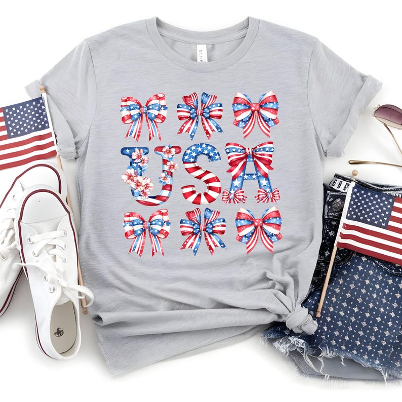 May include: A light gray t-shirt with the word "USA" spelled out with red, white, and blue bows and floral accents. The shirt is styled with white sneakers, denim shorts, and American flags, perfect for patriotic celebrations.