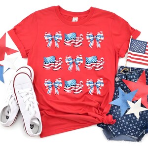 May include: A red t-shirt featuring a patriotic design with bows and American flags. The shirt is paired with white sneakers and denim shorts with star cutouts. An American flag is also present.
