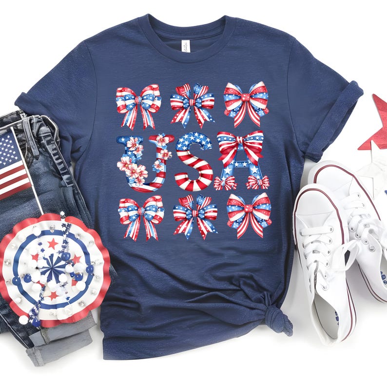 May include: Navy blue t-shirt with a patriotic design. The word "USA" is spelled out with red, white, and blue bows and floral accents. The shirt is paired with denim jeans and white sneakers, perfect for celebrating Independence Day.