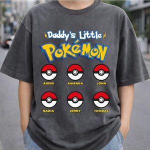 May include: Dark gray t-shirt featuring the text "Daddy's Little Pokémon" in blue and yellow. Below the text are six red and white Poké Ball designs, each with a name: Aiden, Amanda, John, Nadia, Jenny, and Thomas.