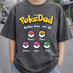 May include: Dark gray t-shirt with the text "PokéDad Gotta love 'em all" in yellow and blue. Below are six colorful Poké Ball designs, each with a name: Robert, Dylan, Johnny, Anna, and Rose. The shirt is a casual style.