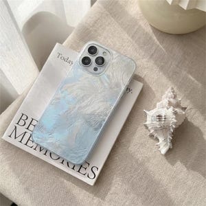 May include: A clear phone case with a pearlescent, textured design. The case is resting on a book with the words "BE MEMORIES" and "Today" visible. A white seashell is next to the book.