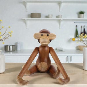 May include: A wooden monkey figurine with a light brown face, ears, and belly, and dark brown body and limbs. The monkey is sitting on a light brown surface with its arms and legs extended. The background is a white kitchen.