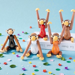 Puede incluir: Cinco figuras de monos de madera en varios colores: negro, blanco, rojo, naranja y marrón. Cada mono tiene brazos y piernas móviles, con un cuerpo marrón claro y detalles amarillos. Los monos están dispuestos sobre una superficie azul claro con flores decorativas.